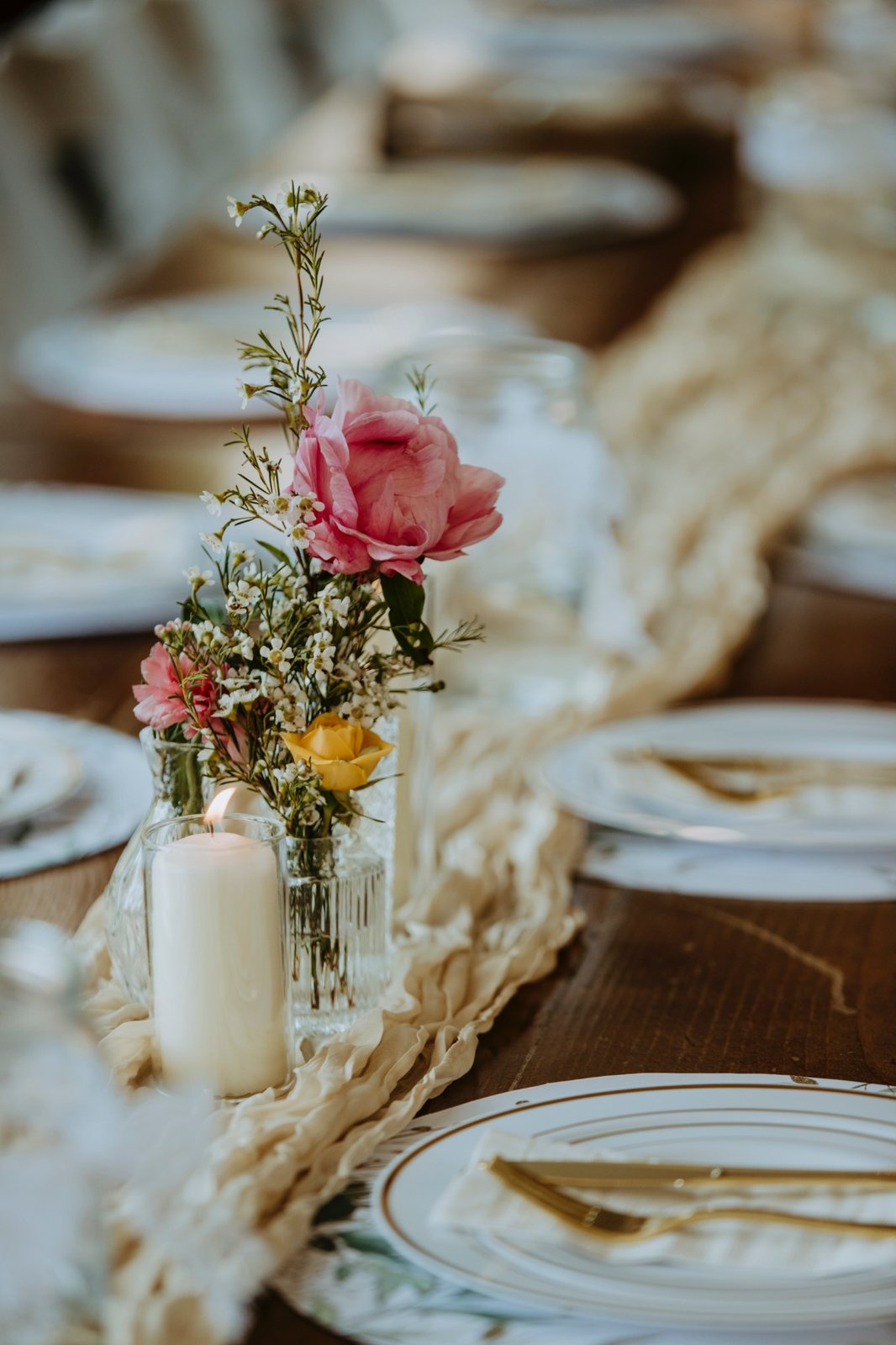Small bud vases with a pink peony, wax flower, and a yellow rose beside a candle
