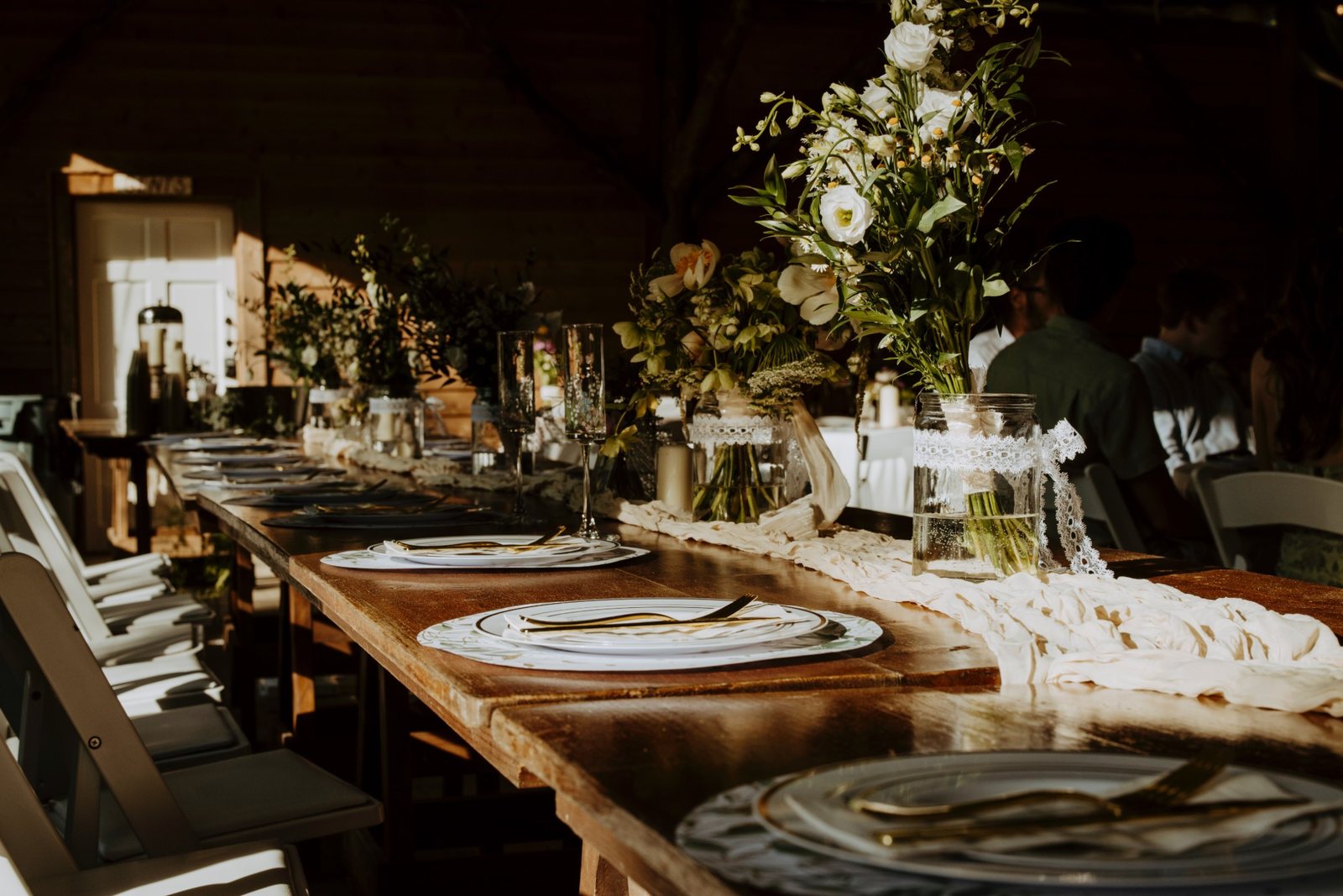 A long dinner table set with bud vases, jars of white flowers, and candles