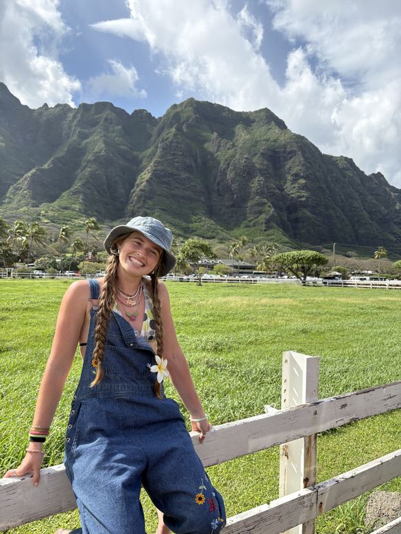 Ria in overalls leaning on a fence, mountains behind her